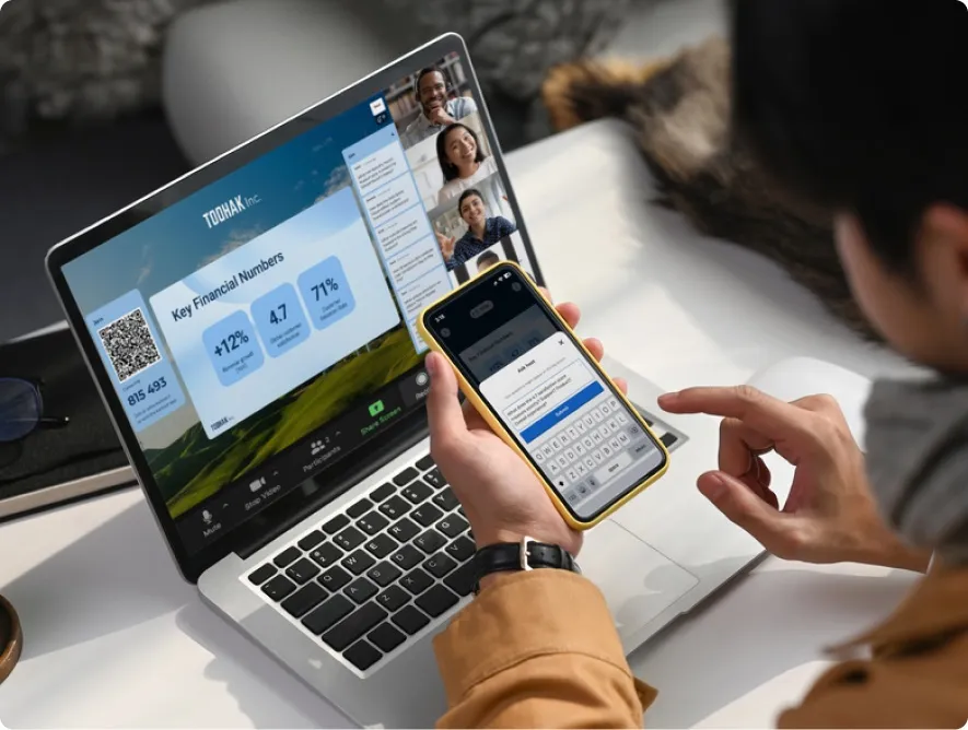 A person participates in a remote meeting on a laptop. The user is also holding a phone to type a question into the "Ask host" feature.